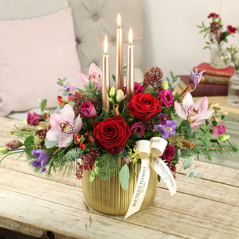 Floral arrangement with candles and a gold vase on a wooden surface, featuring a visible brand name.