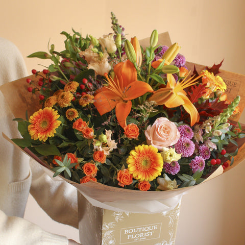 A vibrant autumn floral bouquet wrapped in brown kraft paper and a beige gift box labeled 'BOUTIQUE FLORIST', featuring orange lilies, yellow gerbera daisies, orange roses, purple chrysanthemums, and green foliage.