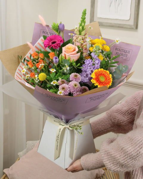 Bouquet of colorful flowers wrapped in paper held by a person wearing a pink sweater.